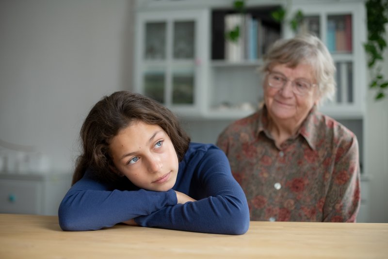 Mädchen mit Oma Ein junge Frau hat den Kopf auf ihre verschränkten Arme gelegt, sie ruhen auf einem Tisch. Hinter ihr sitzt ihre Großmutter und schaut sie nachdenklich an.