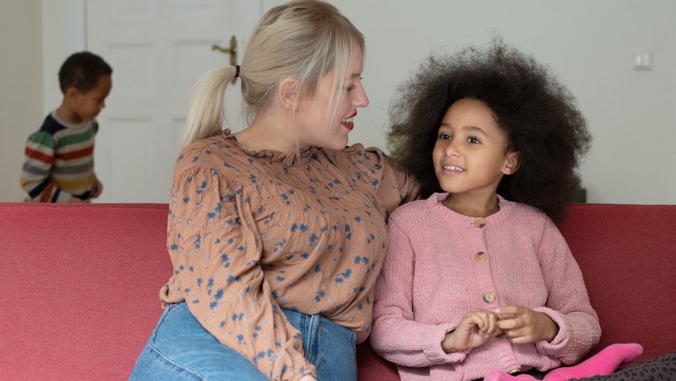 Eine Mutter führt auf dem Sofa ein Gespräch mit ihrer Tochter, im Hintergrund sieht man den kleinen Bruder