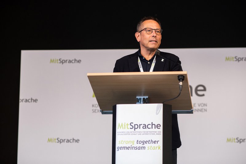 Das Foto zeigt den Unabhängigen Beauftragten Johannes-Wilhelm Rörig, wie er auf dem MitSprache Kongress vom Podium aus eine Ansprache hält. Auf dem Redepult steht "MitSprache" und darunter das Motto des Kongresses "Stronger together – gemeinsam stark".