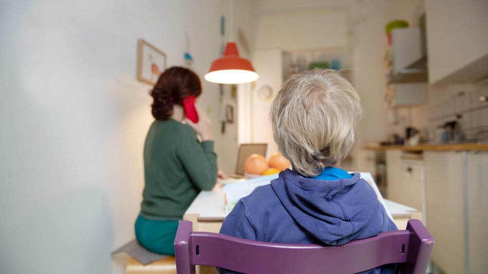 Das Foto wurde in einer Küche aufgenommen: Vor uns sitzt ein Junge, den Rücken zu uns gewandt. Links von ihm sitzt seine Mutter und telefoniert, auch sie wendet sich vom Betrachter ab.