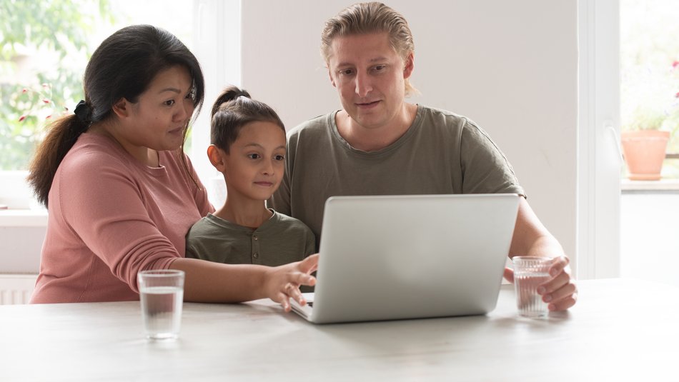 Eine Mutter und ein Vater sitzen vor einem Laptop an einem Tisch. Zwischen ihnen sitzt ihr Kind. Alle schauen auf den Bildschirm.