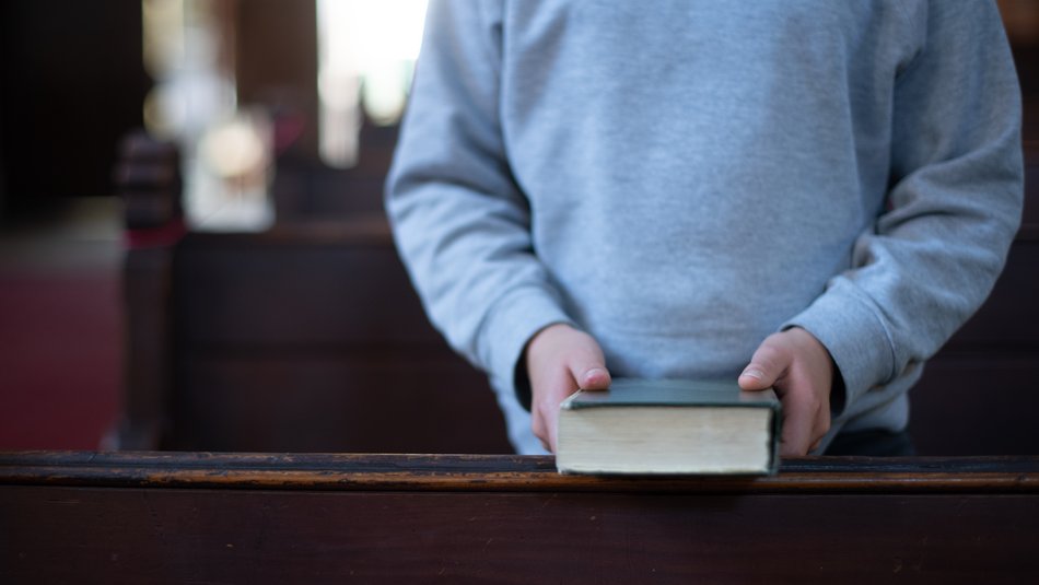 Ein Kind steht mit einer Bibel in der Hand zwischen Kirchenbänken